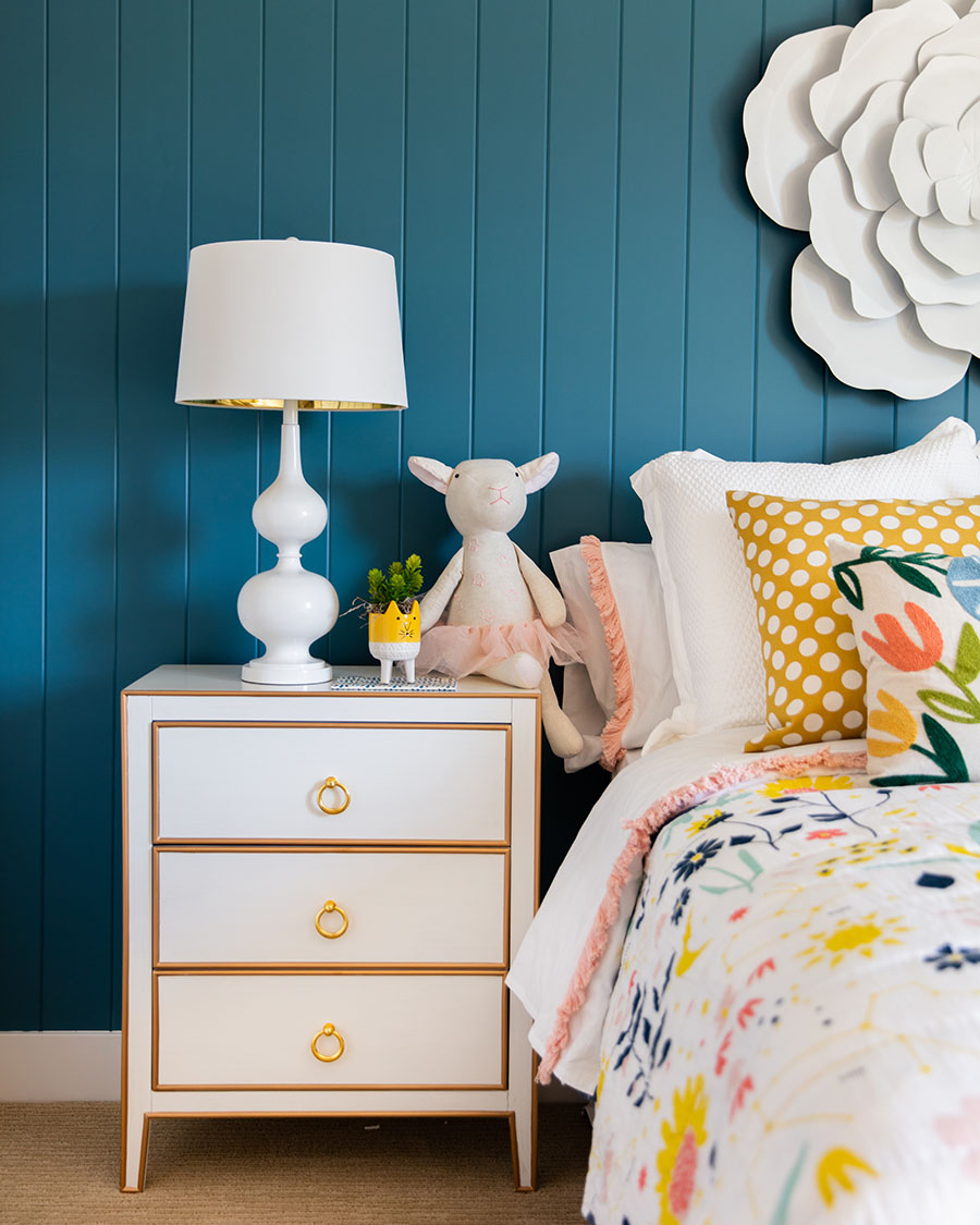 children's bedroom with floral bedding, nightstand with white lamp, blue painted wall and decorative flower on wall